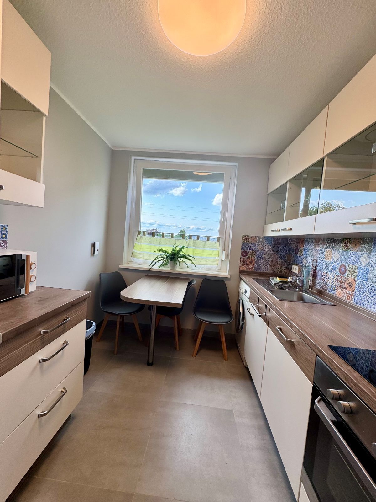 Kleine moderne Küche mit Holzarbeitsplatten, bunten Fliesen, zwei schwarzen Stühlen an einem Holztisch vor einem Fenster mit Blick ins Grüne