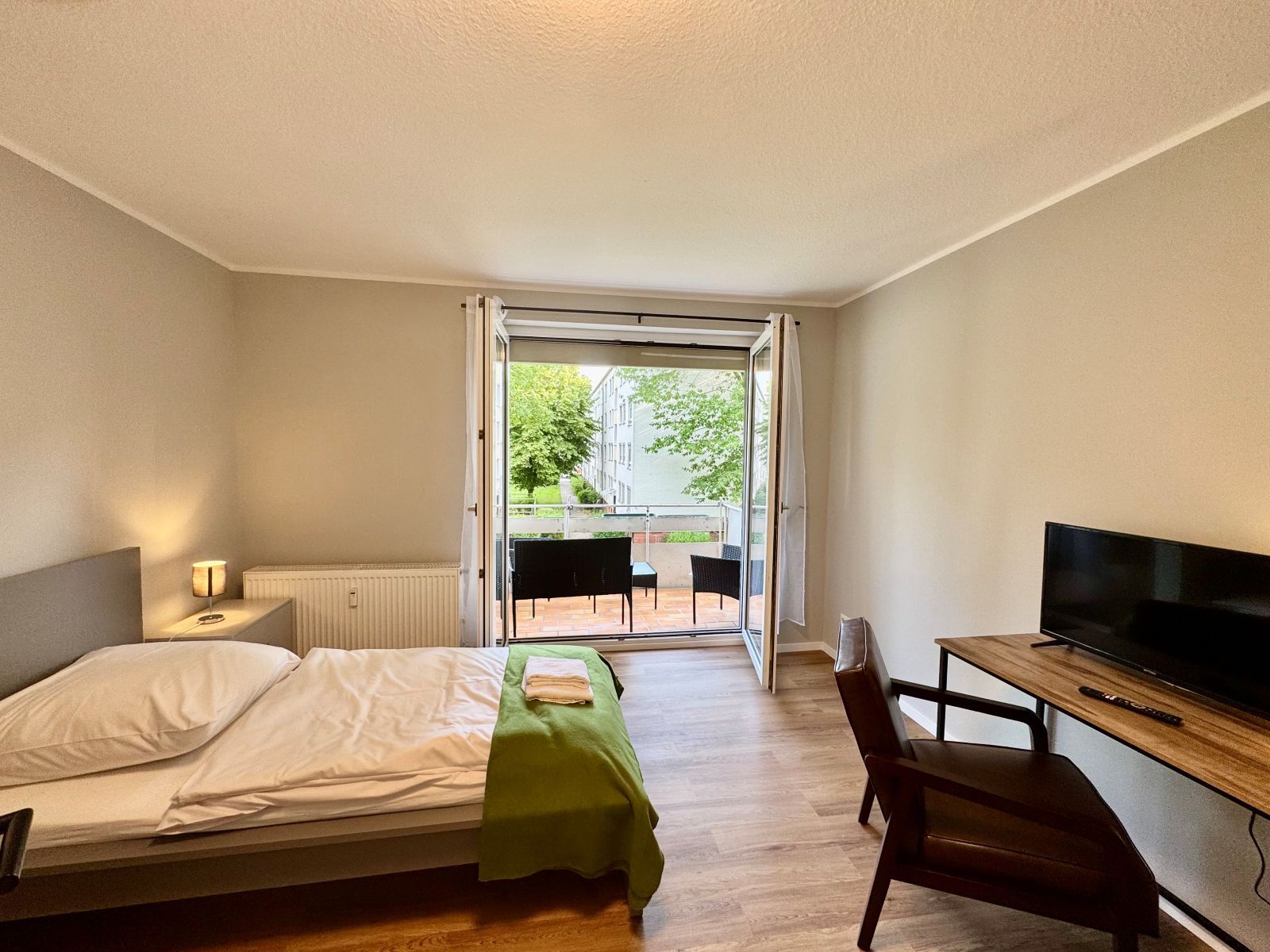 Schlafzimmer mit Einzelbett, grünem Bettüberwurf, Nachttisch mit Lampe, Schreibtisch mit Stuhl und Fernseher, offener Tür zum Balkon mit Blick auf Bäume