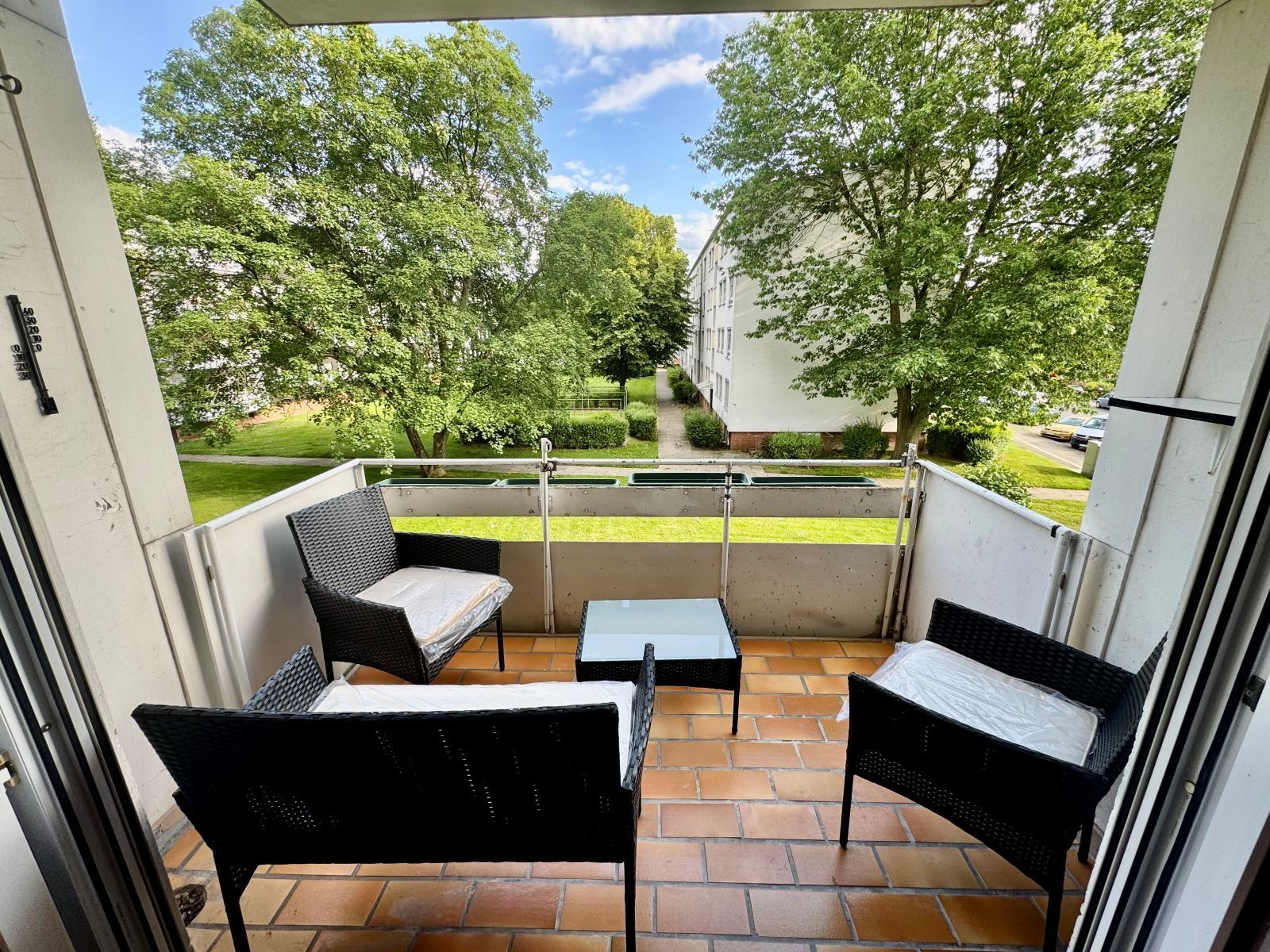 Balkon mit vier schwarzen Stühlen und einem kleinen quadratischen Tisch vor grünen Bäumen und Wohngebäuden