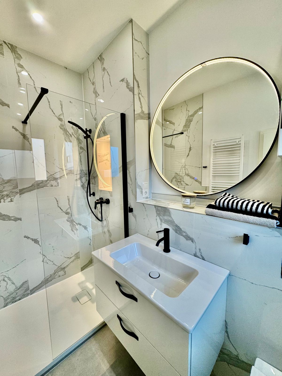 Modernes Badezimmer mit Marmorfliesen, rundem beleuchtetem Spiegel, weißem Waschbecken mit schwarzen Armaturen und begehbarer Dusche mit Glasabtrennung