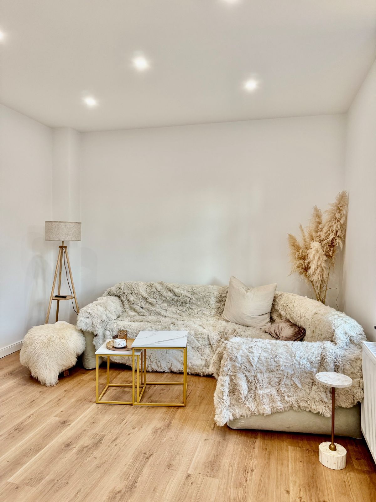 Wohnzimmer mit hellgrauem Ecksofa, flauschigen Decken und zwei kleinen goldfarbenen Beistelltischen