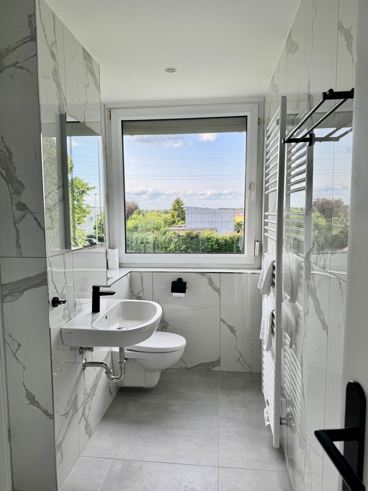 Kleines Badezimmer mit weiß-grauen Marmorfliesen, WC, Wandwaschbecken mit schwarzem Wasserhahn, Handtuchheizkörper und großem Fenster mit Blick ins Grüne