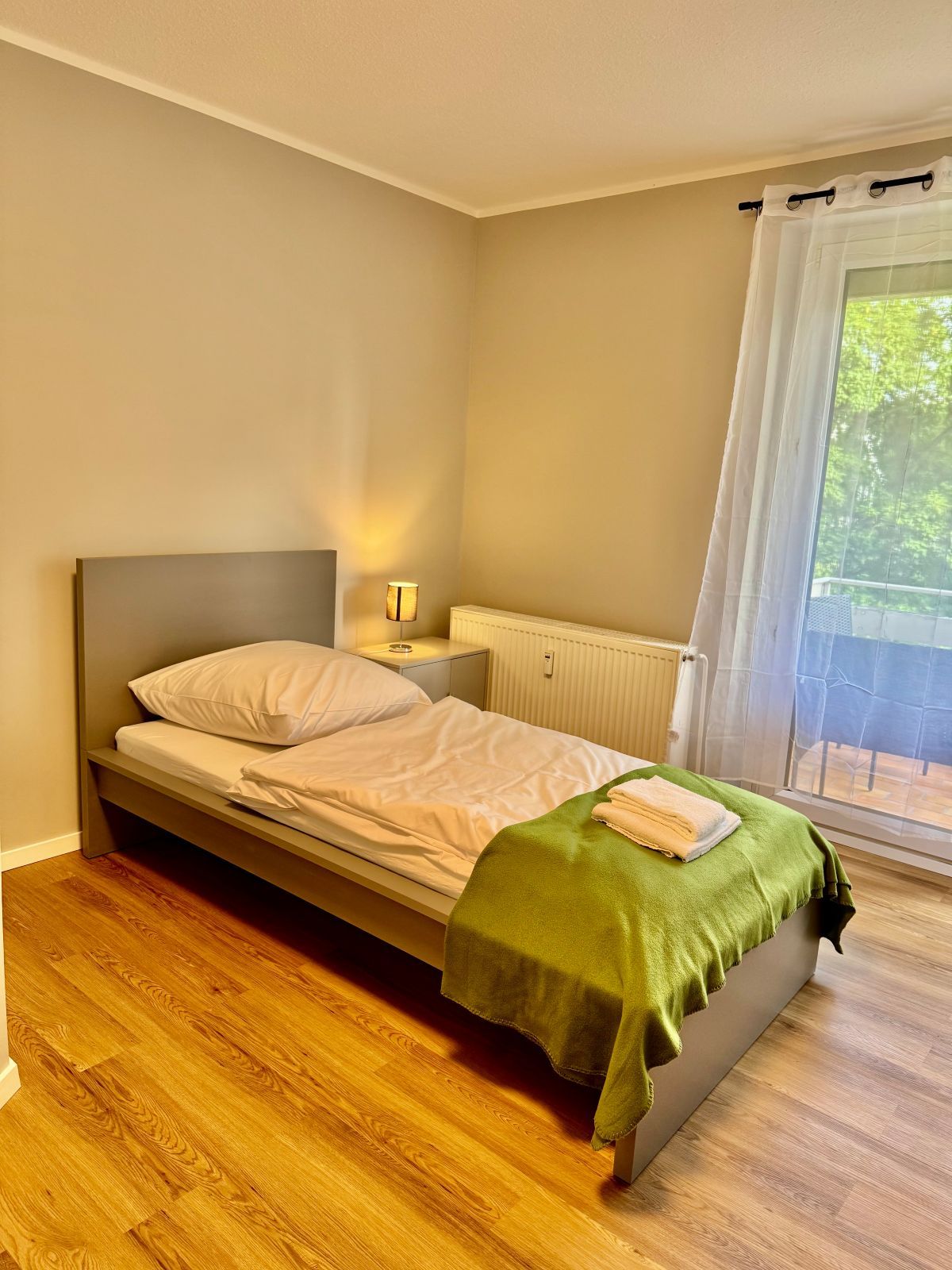 Einzelbett mit grünem Überwurf und weißen Handtüchern in einem hellen Zimmer mit Holzboden und Balkonfenster