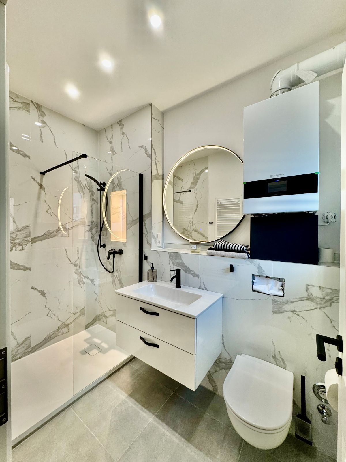 Modernes Badezimmer mit bodengleicher Dusche, rundem beleuchtetem Spiegel, weißem Waschbeckenunterschrank und wandhängendem WC