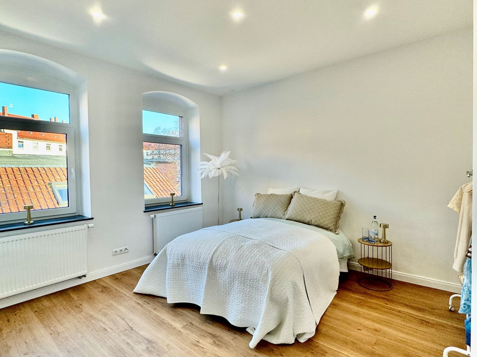 Schlafzimmer mit Doppelbett, weißen Wänden, Holzboden, zwei Fenstern und kleinem runden Nachttisch mit Dekoration